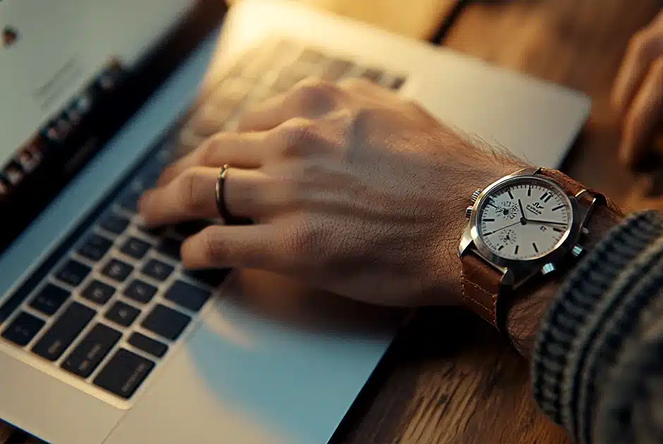 mans arm and hand over a laptop keypad, a watch is on his wrist, plain wooden table surface